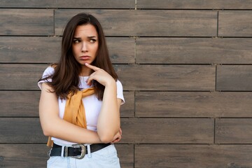 Portrait of touchy sad upset offended resentful young brunet woman wearing casual white t-shirt and jeans with yellow sweater poising near brown wall in the street