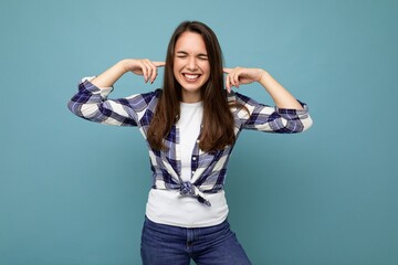 Don't want to hear it. Young emotional positive and smiling attractive brunette woman wearing check shirt isolated on blue background with copy space and covering ears
