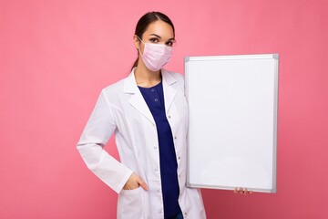 Young attractive nurse in protective face mask holding an empty magnetic board with empty space isolated on pink background