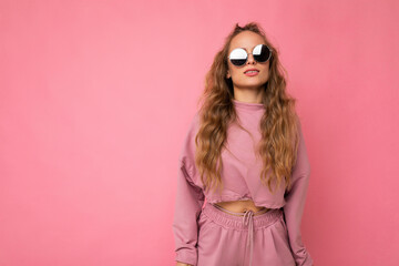 Photo shot of beautiful young dark blonde woman wearing casual clothes and stylish sunglasses isolated over colorful background looking at camera