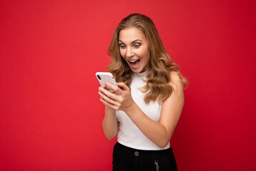 Photo shot of attractive surprised amazed positive good looking young woman wearing casual stylish outfit poising isolated on background with empty space holding in hand and using mobile phone