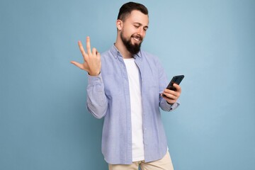 Photo shot of handsome happy smiled satisfued positive good looking young man wearing casual stylish outfit poising isolated on background with empty space holding in hand and using mobile phone