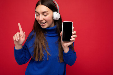 Attractive happy smiling young brunette woman wearing blue sweater isolated over red background holding and using mobile phone surfing on the internet wearing white headsets listening to music looking