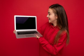 Side profile photo of beautiful happy girl with long hair wearing red hoodie holding computer laptop looking at netbook keyboard and screen monitor isolated over red wall