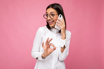 Beautiful happy young brunette woman wearing white blouse and optical glasses standing isolated over pink background talking on mobile phone looking to the side and showing OK gesture