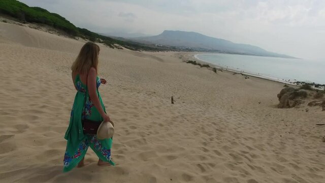 A beautiful girl in a green dress with a hat descends the hill of sand dune to the beach. The sea coast of Tarifa, Spain. The concept of summer holidays. High quality FullHD footage. Slow motion Video