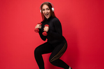 Side-profile photo of beautiful positive smiling young brunette female person wearing black sport clothes isolated on red background wall doing fitness using dumbbells wearing white bluetooth