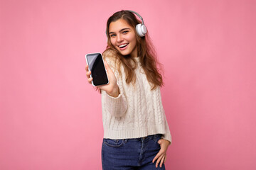 Naklejka premium Beautiful happy smiling young woman wearing stylish casual outfit isolated on background wall holding and showing mobile phone with empty display for mockup wearing white bluetooth headphones