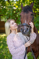 A young rider woman blonde with long hair in a dress posing with brown horse in forest next to farm