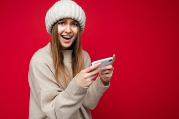 Beautiful emotional happy young blonde woman wearing casual beige sweater and beige hat isolated over red background holding in hand and using mobile phone playing online games looking at camera and