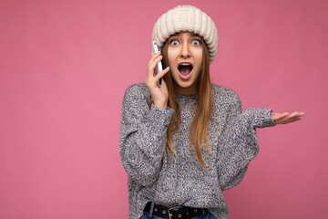 Closeup portrait of Attractive amazed young blonde woman wearing casual grey sweater and beige hat isolated over pink background holding in hand and talking on mobile phone looking at camera with open