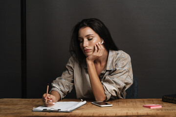 Young brunette woman writing down notes while sitting at desk in office
