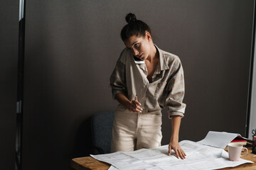 Young brunette woman talking on cellphone while working with drawings