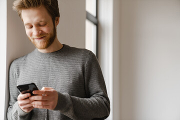 Ginger young man using mobile phone while leaning on wall indoors