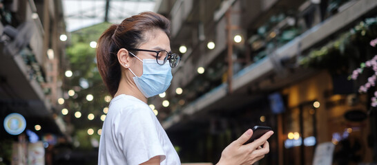 Woman wearing medical face mask and using smartphone during walking in night market, prevent coronavirus or Corona Virus Disease (Covid-19). Health, life concept