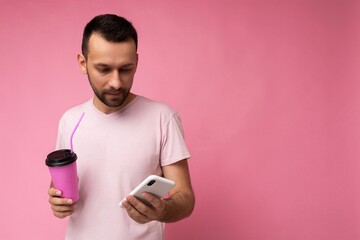 Photo shot of handsome positive good looking young man wearing casual stylish outfit poising isolated on background with empty space holding in hand and using mobile phone messaging sms looking at