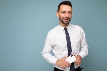 Photo of positive handsome good looking brunet unshaven man with beard wearing casual white shirt and tie isolated on blue background with empty space holding in hand mobile phone looking at camera