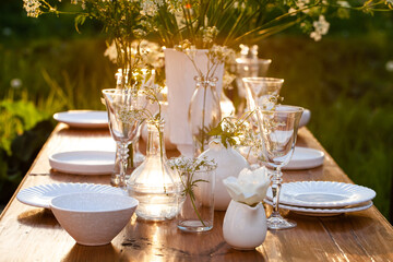 Elegant wedding dinner table decorated with white dishes, flowers in glass vases. Festive atmosphere, luxury party. Sunset, bright summer evening. Close up, details