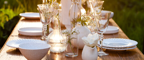 Elegant wedding dinner table decorated with white dishes, flowers in glass vases. Festive atmosphere, luxury party. Sunset, bright summer evening. Close up, details. Banner