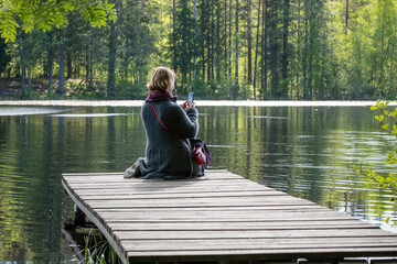A young woman takes photos with a smartphone sitting on the wooden pier on a forest lake on a sunny summer day.