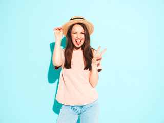 Obraz na płótnie Canvas Portrait of young beautiful smiling female in trendy summer hipster clothes. Sexy carefree woman posing near blue wall in studio. Positive model having fun in hat. Cheerful and happy.Shows peace sign