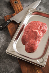 Rinder Steak roh auf Edelstahl Schale mit Küchenmesser, Holzbrett und Stein Hintergrund dunkel