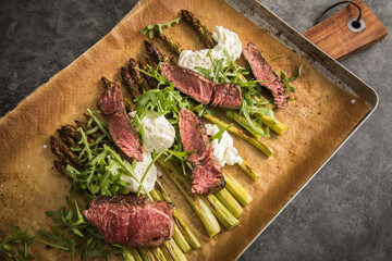 Gerösteter grüner Spargel mit medium rare Rinder Steak Streifen, Rucola, Büffel Mozzarella und Olivenöl auf Backblech mit Backpapier, Holzbrett, Besteck und rustikal dunkel Stein Hintergrund