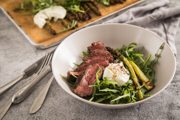 Gerösteter grüner Spargel mit medium rare Rinder Steak Streifen, Rucola, Büffel Mozzarella und Balsamico in Schale und auf Backblech mit Backpapier, Besteck und rustikal Stein Hintergrund
