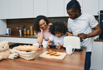 Father, mother and little daughter are preparing food in kitchen of their house. Child looking dish recipe on internet using a smartphone for her family - obrazy, fototapety, plakaty