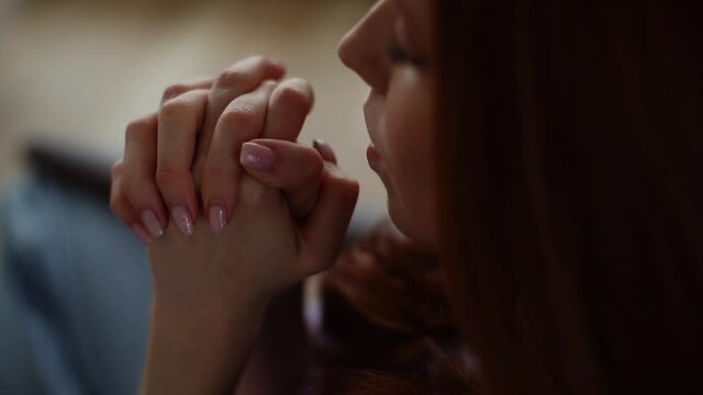 Top close-up view of young woman sincerely praying with eyes closed holding folded hands in front of face at home. Tracking shot of worried female put hands in prayer praying excitedly with hope.