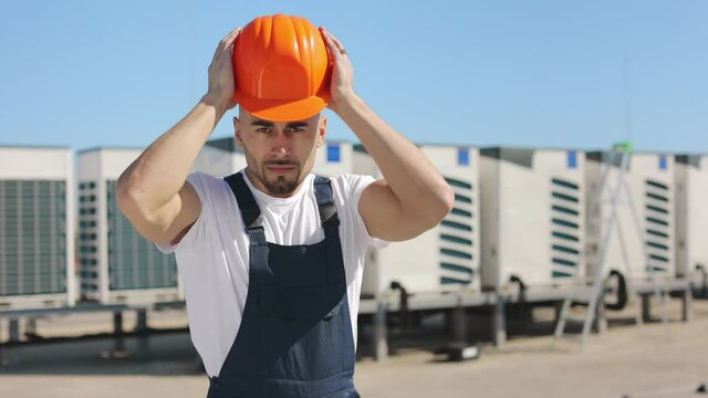 Portrait of a young engineer who is wearing a protective helmet on his head and crossing his arms. He is looking at the camera. He is wearing work clothes. Air conditioning system in the background