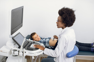 Ultrasound examination of the child in modern clinic. Young afro american female confident doctor makes an abdominal scan of a school 12-aged boy in a medical clinic - Powered by Adobe