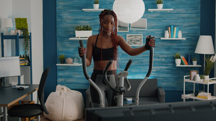 Black woman athletic training on elliptical bike practicing cardio sport watching aerobic video on...