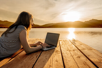 Young woman enjoying her creativeness writing book on a pier at sunset. New job opportunity at modern times to work everywhere using laptop and wifi internet connection technology. Girl studying - obrazy, fototapety, plakaty