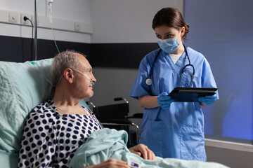 Medical assistant with protection mask checking treatment of senior man reading notes on tablet pc. Sick elderly laying in bed discussing with nurse about diagnosis expertise with iv bag attached.