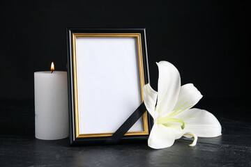 Funeral photo frame with ribbon, white lily and candle on black table against dark background....