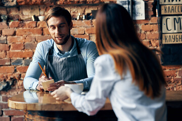 male waiter in gray aprons takes an order and a cup of coffee female client at a table in a cafe