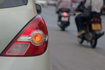 Illuminated turn indicator signal light of on the rear of a white car, blurred on the right are passing motorcycles