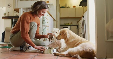 Smiling caucasian woman feeding her pet dogs pouring food into bowl in kitchen at home - Powered by Adobe