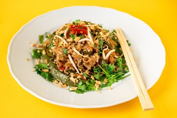 Vietnamese noodle with chicken meat, pea, sprouts, chili coriander herb on white plate, chopstick, on yelllow.