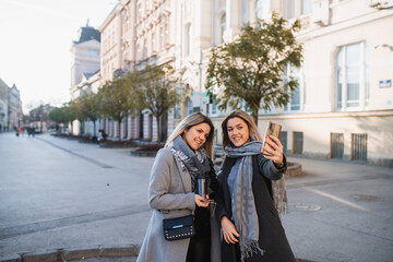 Fototapeta premium Two young women friends take a selfie on the street the background of trees andbuildings looking into the phone in hand and posing.