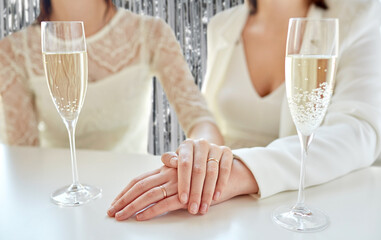 homosexuality, same-sex marriage and lgbt concept - close up of happy married lesbian couple hands with champagne glasses and wedding rings over foil party curtain on background