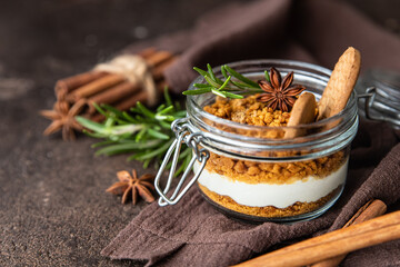 Layered dessert in glass jar with cookie crumble and whipped cream decorated with rosemary and anise, dark brown background. No bake cheesecake, trifle or pudding.