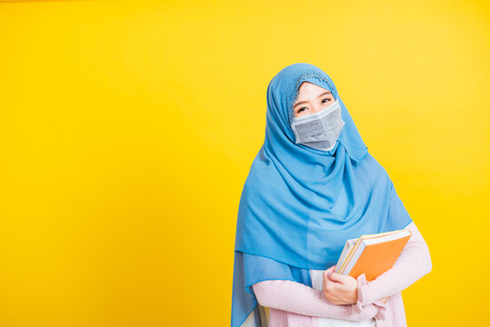 Asian Muslim Arab, Portrait Of Young Woman Religious Wear Veil Hijab And Face Mask Protective To Prevent Coronavirus She Student Hold Books On Hand, Isolated On Yellow Background, Back To College