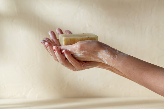 natural cosmetics, hygiene and beauty concept - foamy hands holding bar of craft soap on beige background
