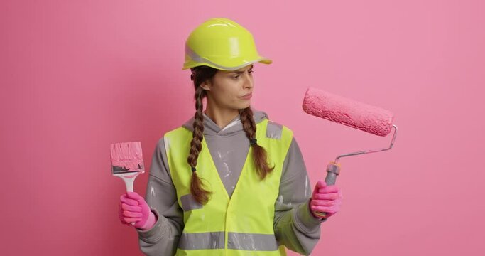 Puzzled woman repairer holds painting roller and brush busy doing home makeover uses repair tools wears helmet safety uniform isolated over pink background. Maintenance service and remodeling