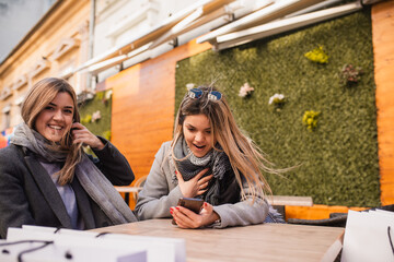 Two young blonde women, 25 years old, are sitting in a cafe, chatting and looking at the phone. Fashion and influential
