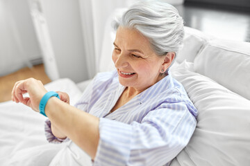 old age, technology and people concept - happy smiling senior woman in pajamas with health tracker sitting in bed at home bedroom