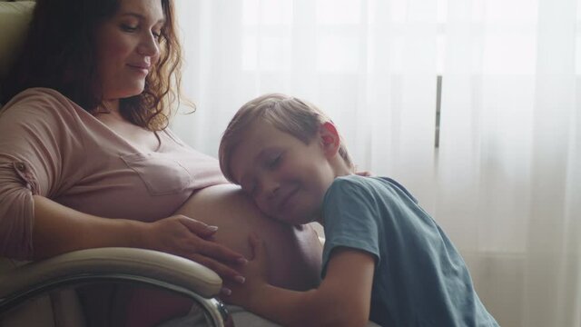Child hugs and kisses her mother's pregnant belly. Happy son strokes the big belly his pregnant mom near window at home. Happy family life in an expectation a newborn. Pregnancy and maternity.