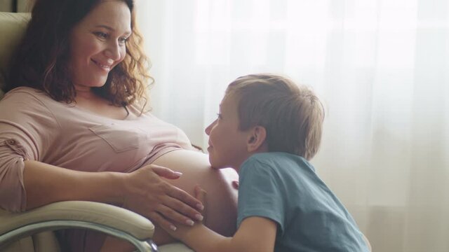 Child hugs her mother's pregnant belly. Happy son strokes the big belly his pregnant mom near window at home. Happy family life in an expectation a newborn. Pregnancy and maternity.